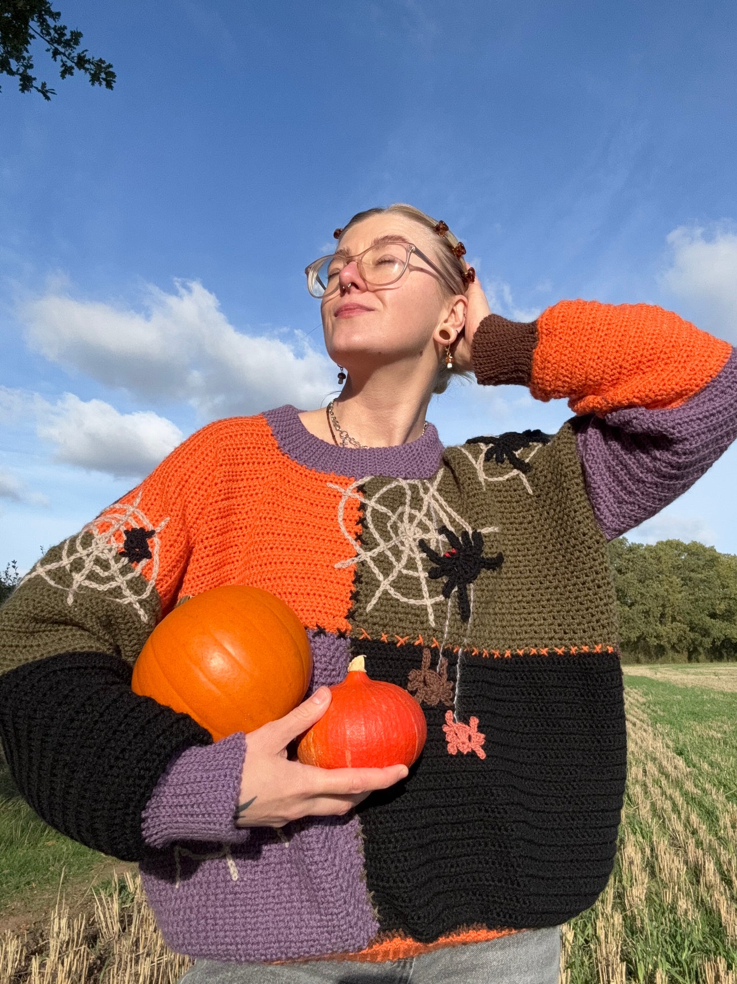 Beginner-friendly autumn patchwork jumper crochet pattern, cozy fit for fall season. Sweater in green, brown, orange, purple colors. Model wearing chunky autumn sweater at pumpkin patch, styled for October weather.